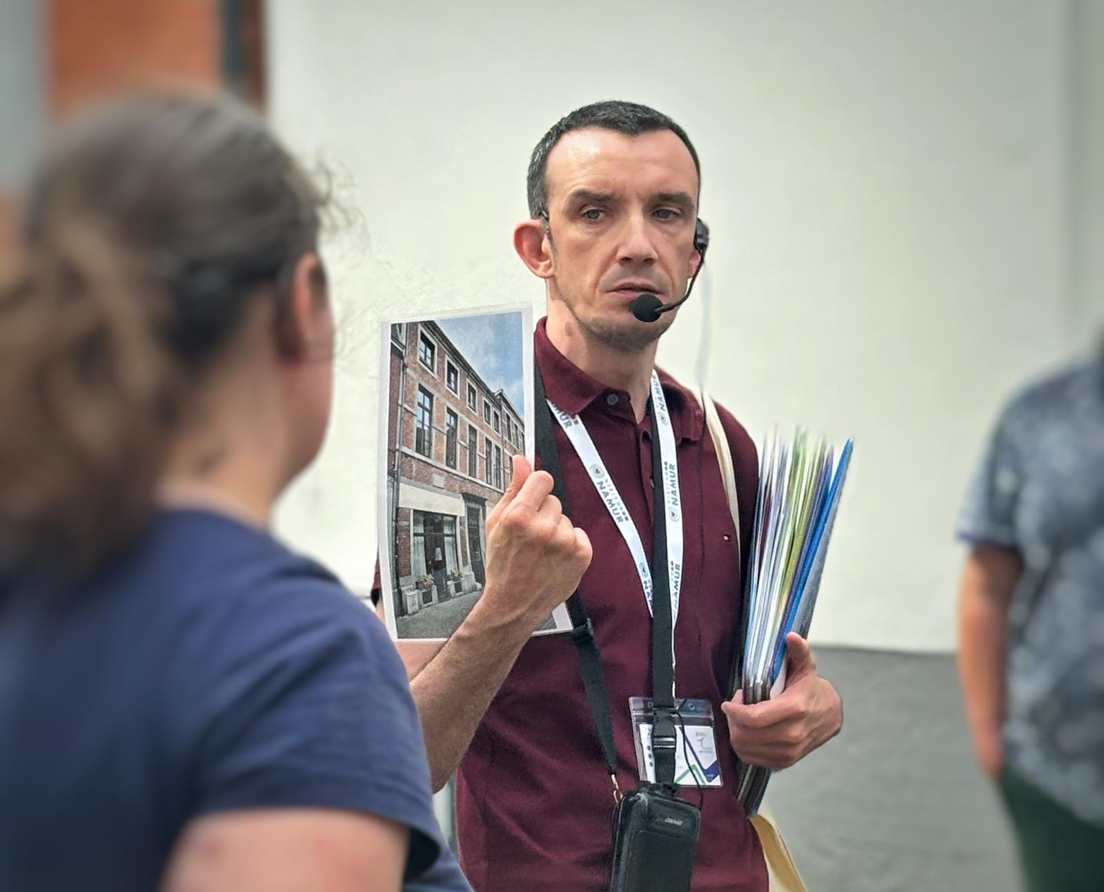 ALT: guide touristique de face qui montre une image. Il a un micro casque, une farde et un badge au cou. On voit une dame et un homme de dos.